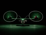 Anduril unveils armed drone gunship to fly with Apaches Front view of a futuristic dual-rotor aircraft with two large glowing green circular rotors, parked in a dark hangar.
