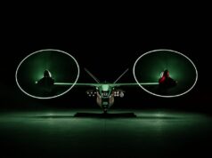 Anduril unveils armed drone gunship to fly with Apaches Front view of a futuristic dual-rotor aircraft with two large glowing green circular rotors, parked in a dark hangar.