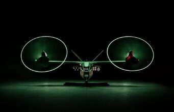 Anduril unveils armed drone gunship to fly with Apaches Front view of a futuristic dual-rotor aircraft with two large glowing green circular rotors, parked in a dark hangar.