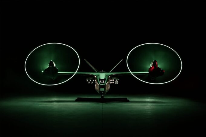 Anduril unveils armed drone gunship to fly with Apaches Front view of a futuristic dual-rotor aircraft with two large glowing green circular rotors, parked in a dark hangar.