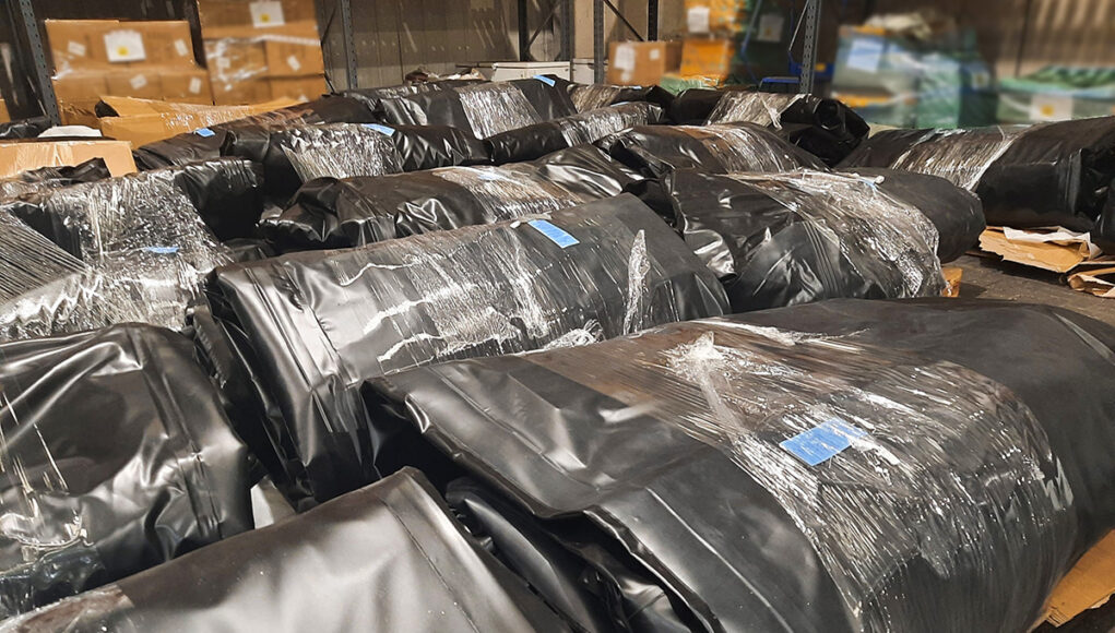 Warehouse scene with large black plastic-wrapped bundles on pallets, stacked in aisles.