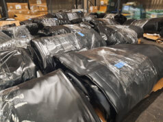 Warehouse scene with large black plastic-wrapped bundles on pallets, stacked in aisles.