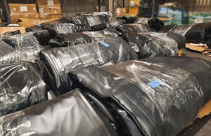 Warehouse scene with large black plastic-wrapped bundles on pallets, stacked in aisles.