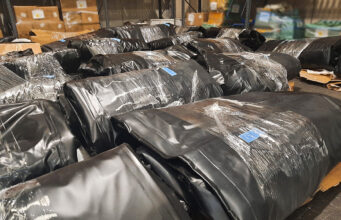 NCA seizes 25 smuggling boats and engines at Felixstowe Warehouse scene with large black plastic-wrapped bundles on pallets, stacked in aisles.