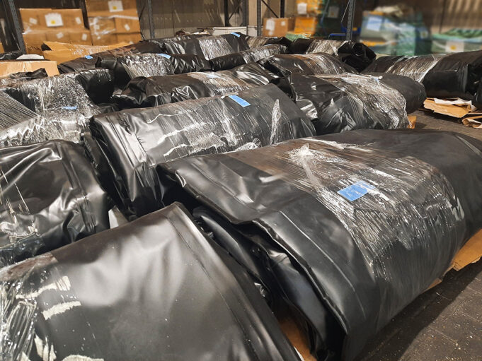 Warehouse scene with large black plastic-wrapped bundles on pallets, stacked in aisles.