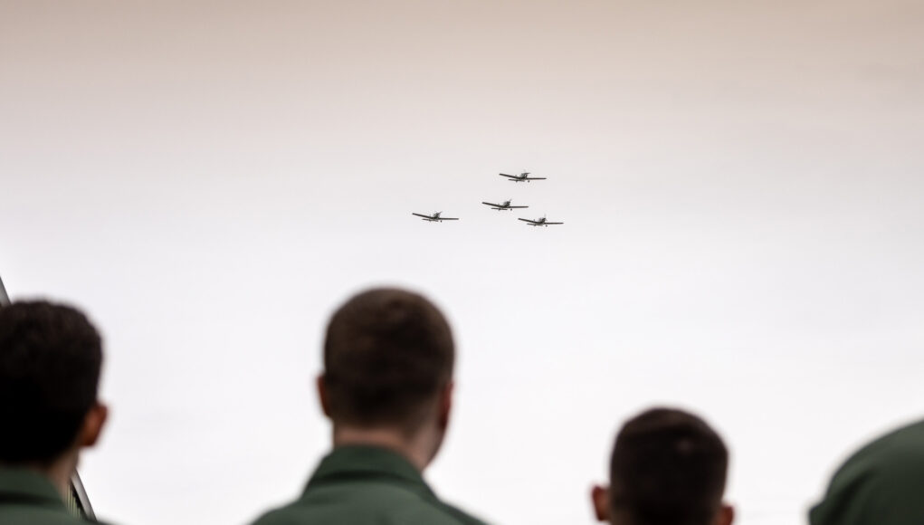 People in green uniforms seen from behind watch four small planes in tight formation flying overhead?