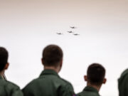 People in green uniforms seen from behind watch four small planes in tight formation flying overhead?