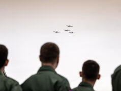 People in green uniforms seen from behind watch four small planes in tight formation flying overhead?