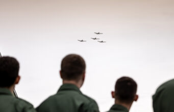 People in green uniforms seen from behind watch four small planes in tight formation flying overhead?