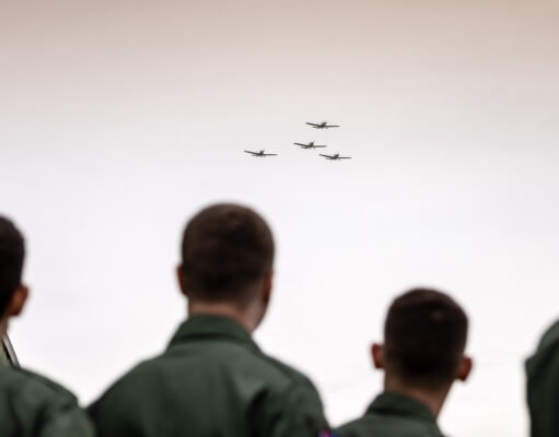 People in green uniforms seen from behind watch four small planes in tight formation flying overhead?