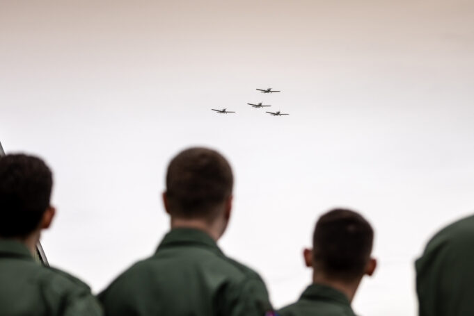 People in green uniforms seen from behind watch four small planes in tight formation flying overhead?