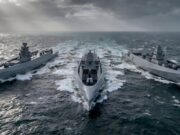 Three modern gray warships sail in rough seas, creating white wake under a stormy, sun-slashed sky.