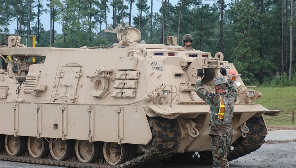 Beige armored tracked vehicle on a road; a soldier stands on top while another signals at the front, in a wooded area behind.
