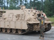 Beige armored tracked vehicle on a road; a soldier stands on top while another signals at the front, in a wooded area behind.