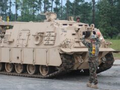 Beige armored tracked vehicle on a road; a soldier stands on top while another signals at the front, in a wooded area behind.