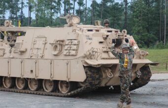 BAE signs Polish deal towards local armoured vehicle build Beige armored tracked vehicle on a road; a soldier stands on top while another signals at the front, in a wooded area behind.