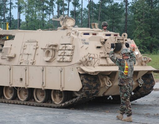 Beige armored tracked vehicle on a road; a soldier stands on top while another signals at the front, in a wooded area behind.