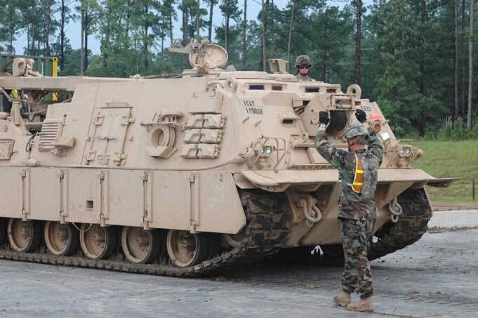 BAE signs Polish deal towards local armoured vehicle build Beige armored tracked vehicle on a road; a soldier stands on top while another signals at the front, in a wooded area behind.