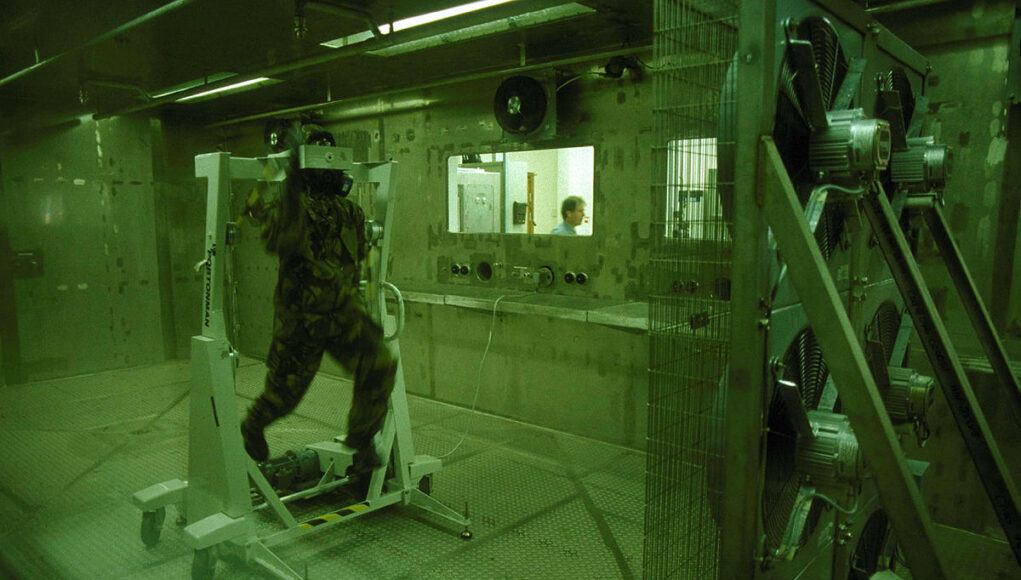 Soldier in camouflage gear walking on a treadmill inside a sealed lab chamber, with a technician visible through a window in the background.