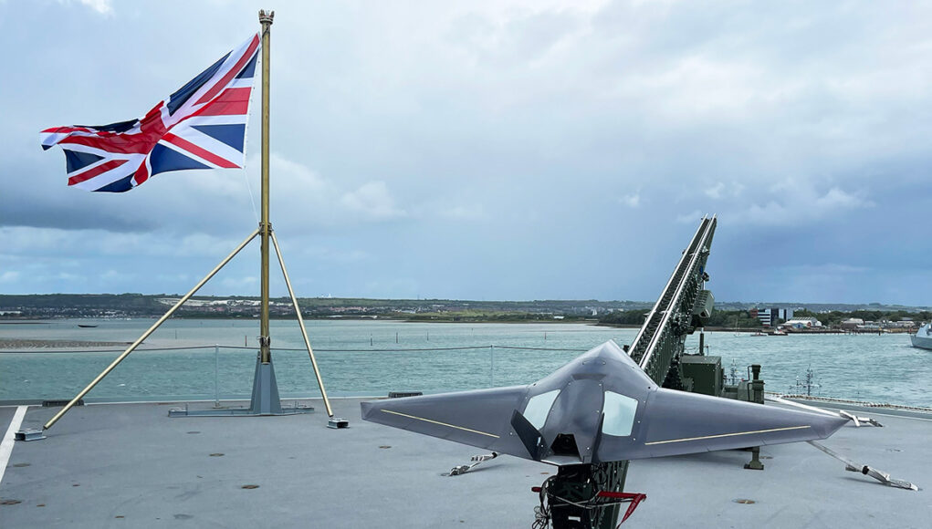 RS290623_IMG_3975 Union Jack flag flies on a naval deck with a gray stealth jet on a launch catapult overlooking the harbor