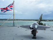 Strike drone launched from Royal Navy ship for first time Union Jack flag flies on a naval deck with a gray stealth jet on a launch catapult overlooking the harbor