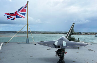 Union Jack flag flies on a naval deck with a gray stealth jet on a launch catapult overlooking the harbor