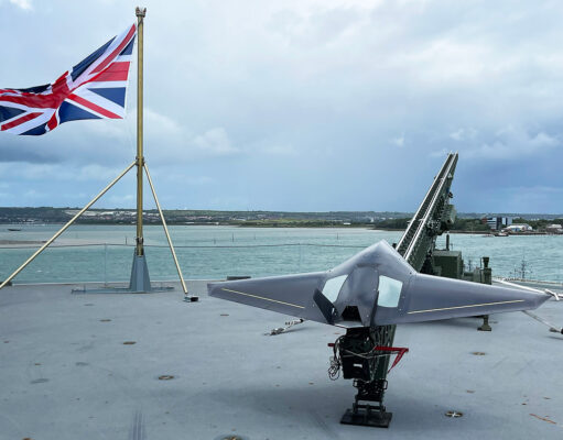 Strike drone launched from Royal Navy ship for first time Union Jack flag flies on a naval deck with a gray stealth jet on a launch catapult overlooking the harbor