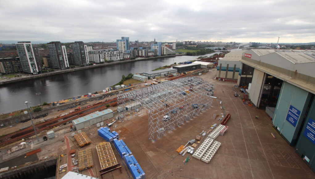 RS293542_Govan paint facility under construction 13 July_scr Industrial shipyard scene: a large white metal frame under construction beside a riverside, with stacked blue crates, pallets, and a nearby warehouse and cranes; city buildings in the distance across the water.