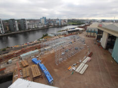 Glasgow warship yard begins work on new facility Industrial shipyard scene: a large white metal frame under construction beside a riverside, with stacked blue crates, pallets, and a nearby warehouse and cranes; city buildings in the distance across the water.