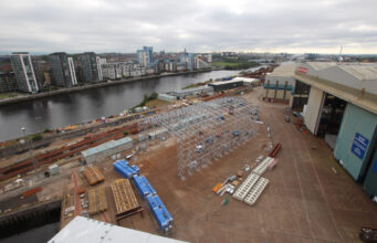 Glasgow warship yard begins work on new facility Industrial shipyard scene: a large white metal frame under construction beside a riverside, with stacked blue crates, pallets, and a nearby warehouse and cranes; city buildings in the distance across the water.