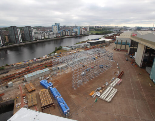 Glasgow warship yard begins work on new facility Industrial shipyard scene: a large white metal frame under construction beside a riverside, with stacked blue crates, pallets, and a nearby warehouse and cranes; city buildings in the distance across the water.
