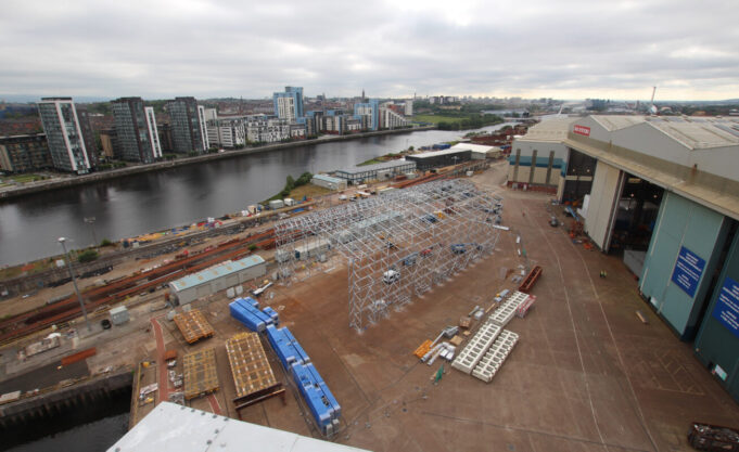 Glasgow warship yard begins work on new facility Industrial shipyard scene: a large white metal frame under construction beside a riverside, with stacked blue crates, pallets, and a nearby warehouse and cranes; city buildings in the distance across the water.