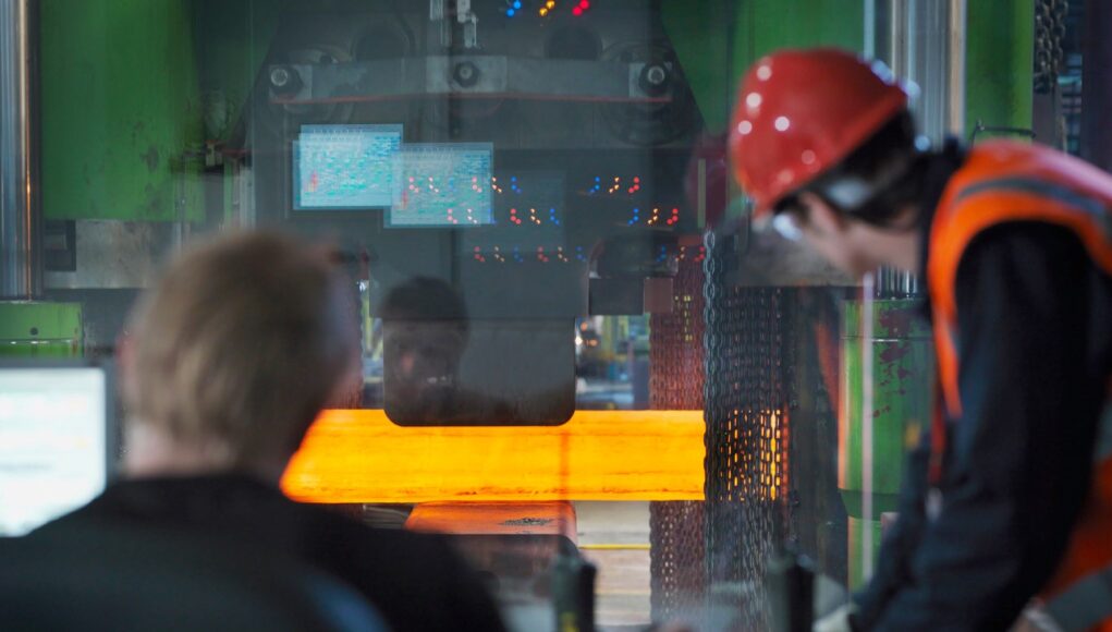 RS294076_GunBarrels226 Two workers in hard hats and safety vests monitor a large metal-forming machine as an orange-hot metal bar passes through it in an industrial setting.
