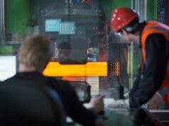 Two workers in hard hats and safety vests monitor a large metal-forming machine as an orange-hot metal bar passes through it in an industrial setting.