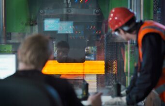 Two workers in hard hats and safety vests monitor a large metal-forming machine as an orange-hot metal bar passes through it in an industrial setting.