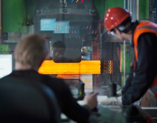 Two workers in hard hats and safety vests monitor a large metal-forming machine as an orange-hot metal bar passes through it in an industrial setting.