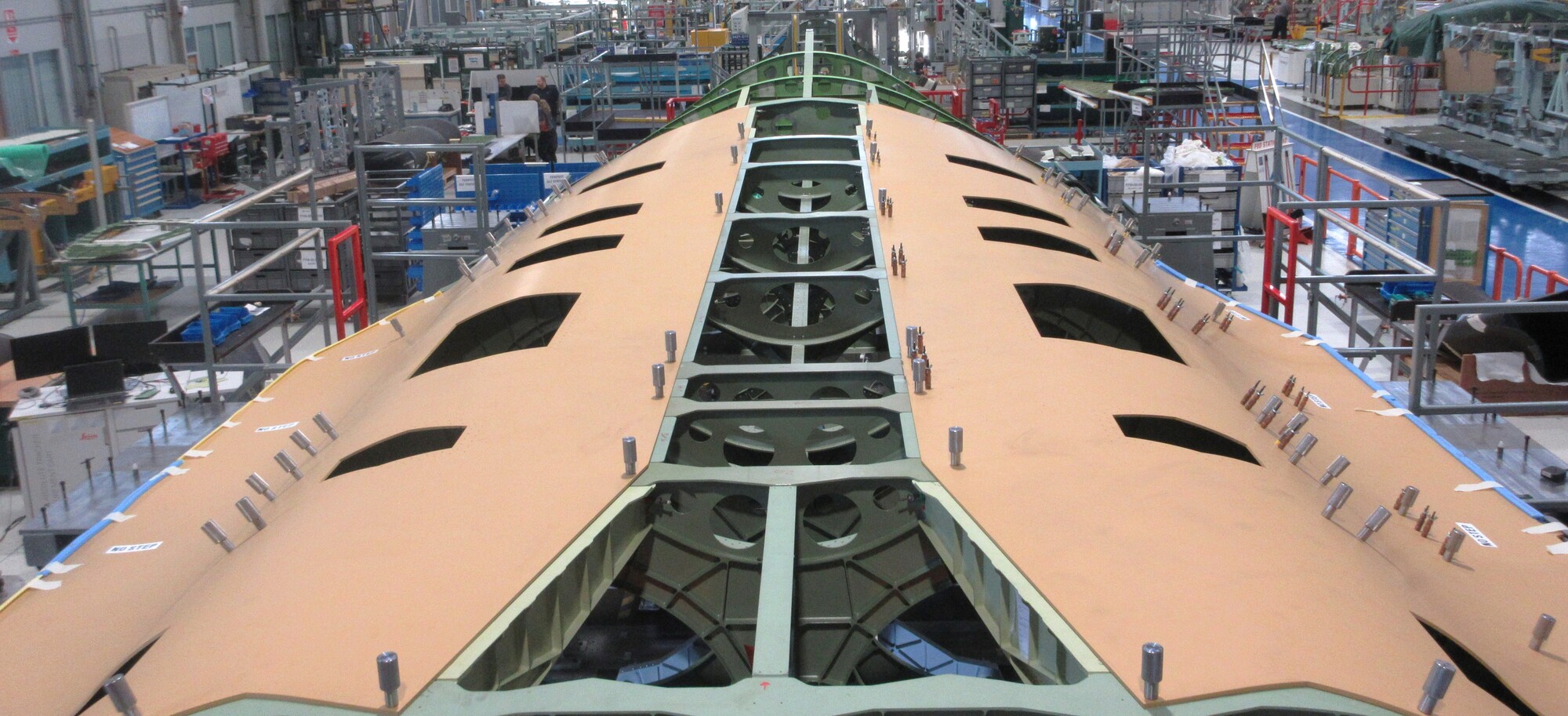 Large aircraft fuselage section under construction in a busy manufacturing hangar, with tools, scaffolding, and workers in the background.