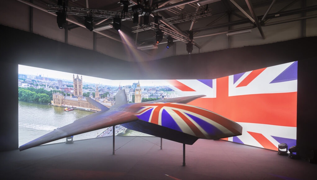 British-themed aircraft model on display in a dark exhibition space, with a curved screen showing London landmarks and the Union Jack backdrop.