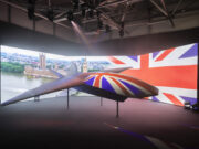 BAE unveils Brontanax, first sovereign UK fighter drone British-themed aircraft model on display in a dark exhibition space, with a curved screen showing London landmarks and the Union Jack backdrop.
