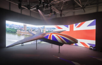BAE unveils Brontanax, first sovereign UK fighter drone British-themed aircraft model on display in a dark exhibition space, with a curved screen showing London landmarks and the Union Jack backdrop.