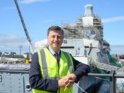 Man in a neon safety vest leaning on a railing at a shipyard, with a scaffolding-covered vessel in the background.