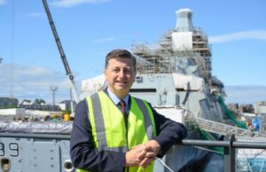 Clyde shipbuilding future backed on Glasgow yard visit Man in a neon safety vest leaning on a railing at a shipyard, with a scaffolding-covered vessel in the background.