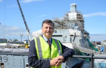 Clyde shipbuilding future backed on Glasgow yard visit Man in a neon safety vest leaning on a railing at a shipyard, with a scaffolding-covered vessel in the background.