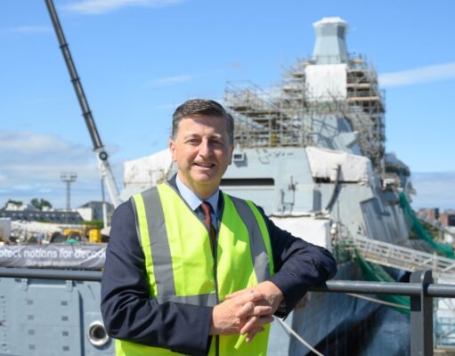 Clyde shipbuilding future backed on Glasgow yard visit Man in a neon safety vest leaning on a railing at a shipyard, with a scaffolding-covered vessel in the background.