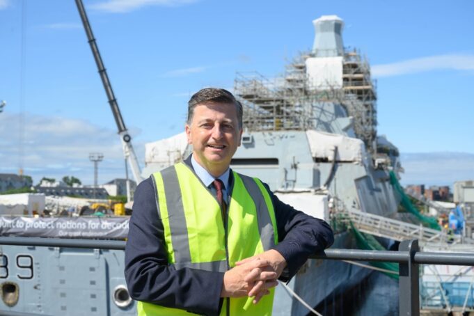 Man in a neon safety vest leaning on a railing at a shipyard, with a scaffolding-covered vessel in the background.