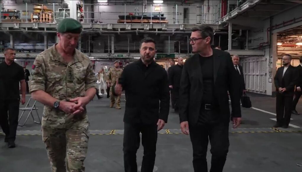 Three men walk through a large industrial hangar: a soldier in camouflage with a green beret, flanked by two men in black outfits, with others in the background.