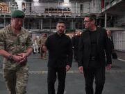Three men walk through a large industrial hangar: a soldier in camouflage with a green beret, flanked by two men in black outfits, with others in the background.