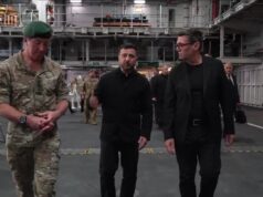 Three men walk through a large industrial hangar: a soldier in camouflage with a green beret, flanked by two men in black outfits, with others in the background.