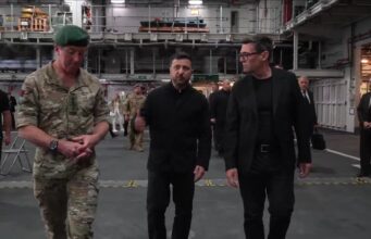 Three men walk through a large industrial hangar: a soldier in camouflage with a green beret, flanked by two men in black outfits, with others in the background.
