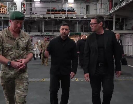 Three men walk through a large industrial hangar: a soldier in camouflage with a green beret, flanked by two men in black outfits, with others in the background.